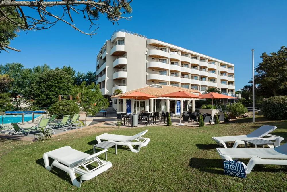 Hôtel Valdys Thalasso & Spa Les Pins à Saint-Jean-de-Monts : bâtiment blanc à étages, piscine extérieure et transats sur la pelouse sous les pins