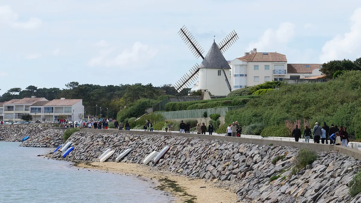 Le moulin de la Conchette dominant le front de mer de Jard-sur-Mer, avec des promeneurs sur la digue et des barques échouées
