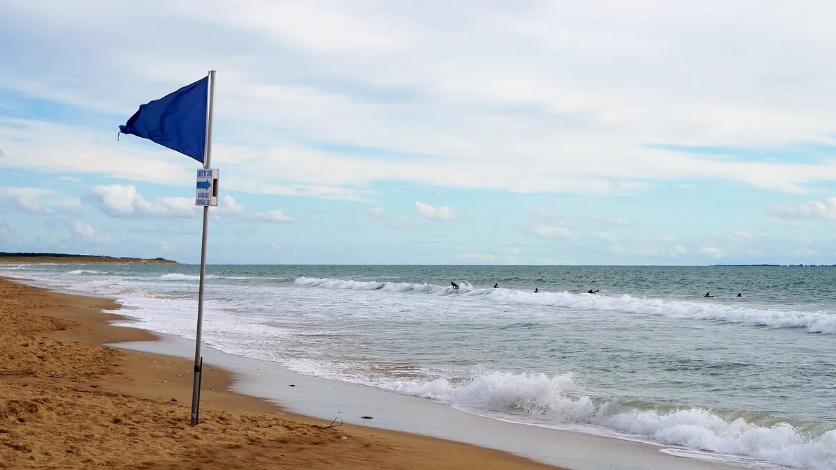 Vagues, drapeau bleu et sable de la plage des Conches à Longeville-sur-Mer, sous un ciel nuageux