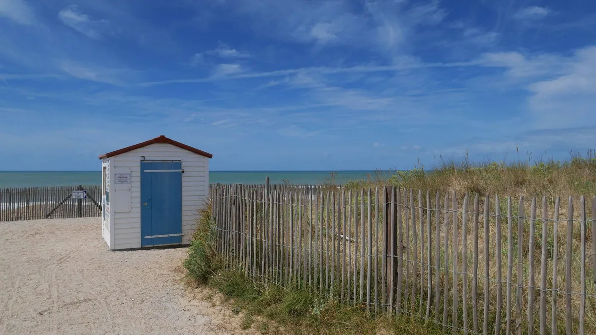 Cabane blanche au toit rouge et à la porte bleue entre les dunes et l'océan, sur une plage de Saint-Hilaire-de-Riez