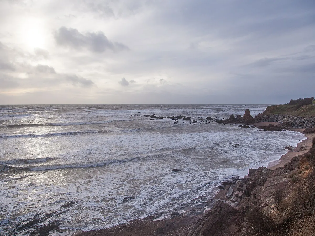 Côte de la Sauzaie à Brétignolles-sur-Mer par temps agité : vagues sur une petite plage de sable bordée de rochers, sous un ciel nuageux