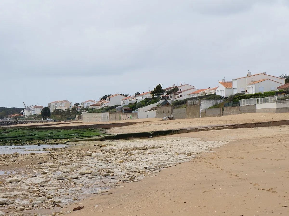Plage de sable et de rochers à Jard-sur-Mer, avec des maisons blanches sur le front de mer sous un ciel nuageux