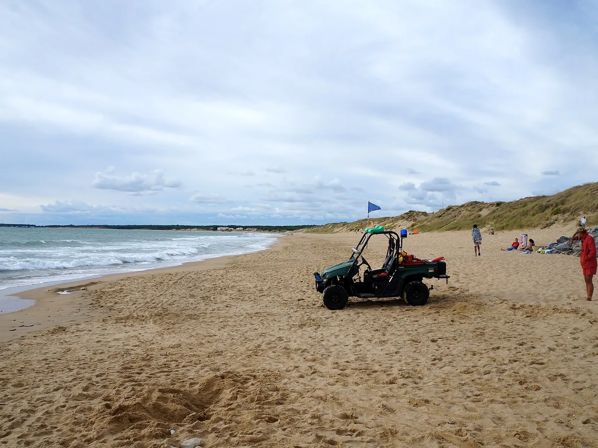 La plage de sable des Conches à Longeville-sur-Mer, avec un petit véhicule vert, un drapeau bleu et des dunes à l'arrière-plan