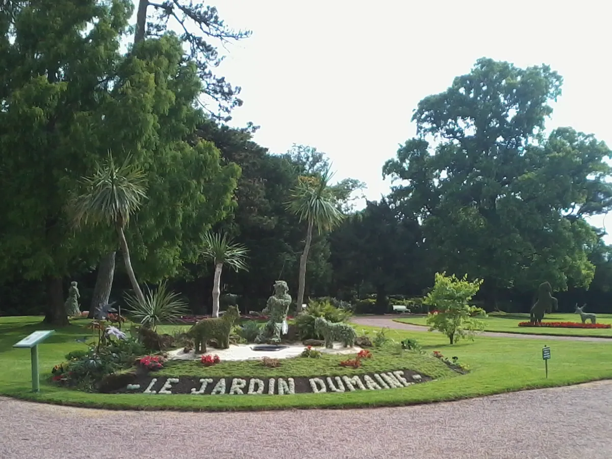 Pelouse du jardin Dumaine à Luçon, avec un parterre fleuri portant l'inscription « Le Jardin Dumaine », des topiaires et de grands arbres