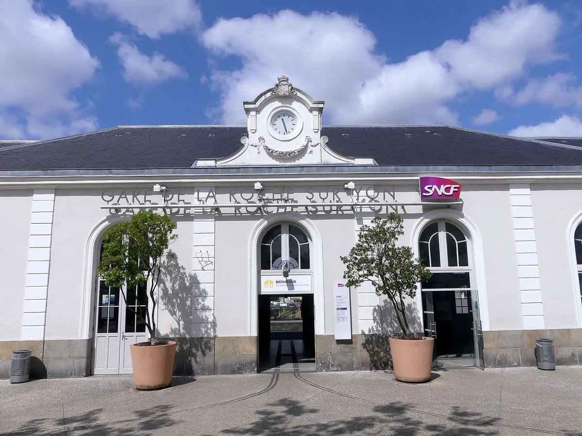 Façade blanche de la gare de La Roche-sur-Yon, avec son horloge, son inscription et deux grands pots d'arbustes devant l'entrée